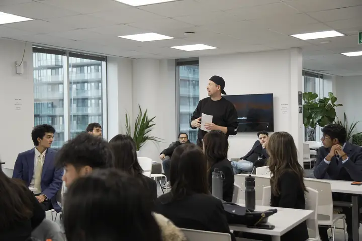 A man in a black cap standing with notes in his hand, speaking to a room of seated students.