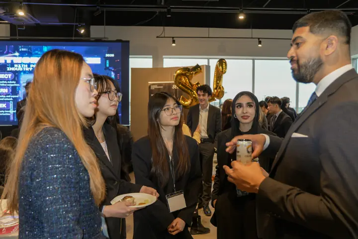A man in a suit talking with a group of students at LUMEN 2026, gold balloons behind them.