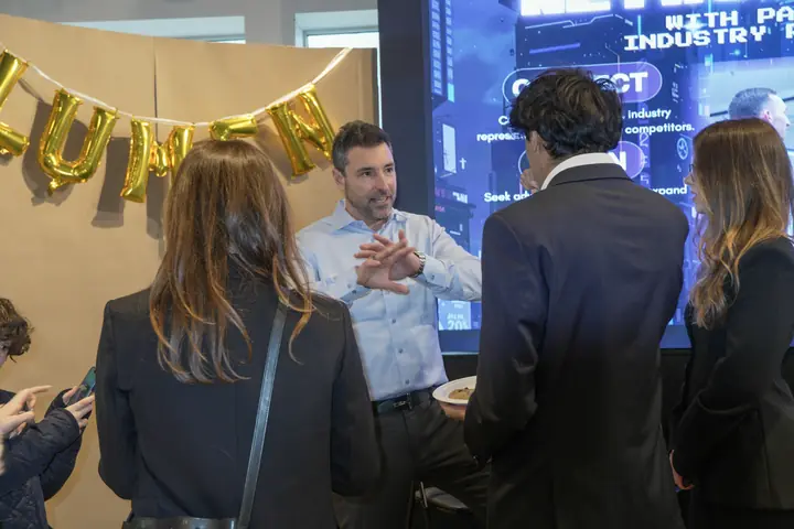 A man in a blue shirt gesturing as he talks with three students, gold balloons spelling LUMEN behind them.
