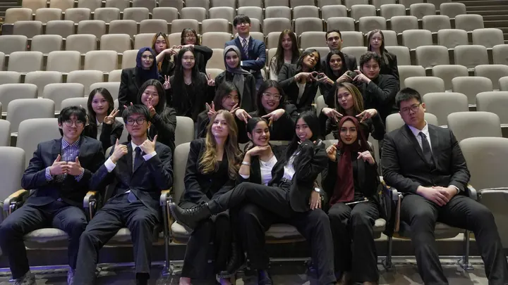 The SEC team: a large group in dark suits seated together in a lecture hall, making heart shapes with their hands.