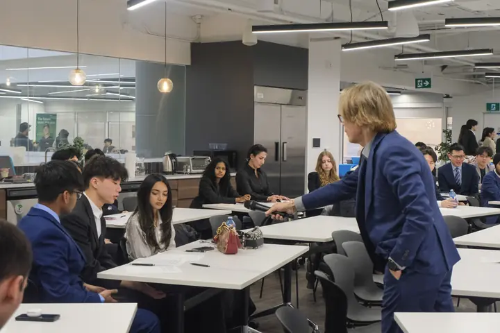 A man in a blue suit holds a microphone out to students seated at long white desks, at the UEC 2025 workshop.